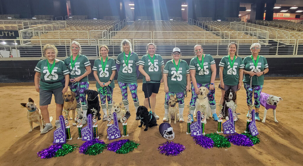 Ten women in football jersey's standing with their dogs with trophies and pom poms in front of them